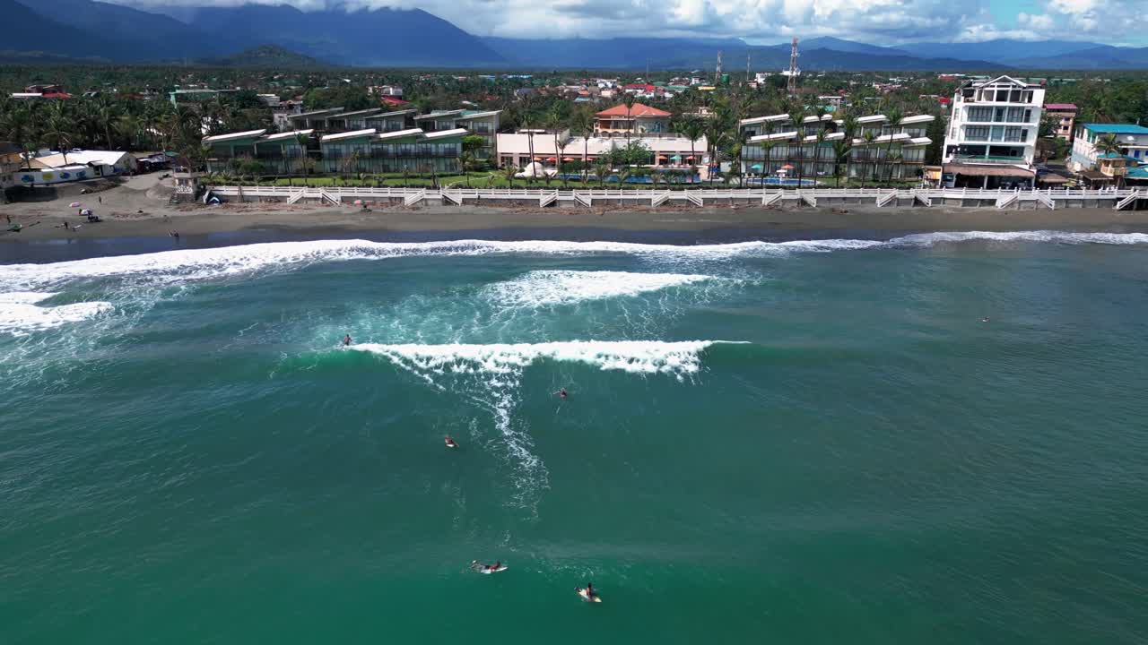 A dolly-out wide shot gracefully pulls back from the shore, revealing surfers riding the waves along the coastline, while beach houses are scattered across the sandy expanse.