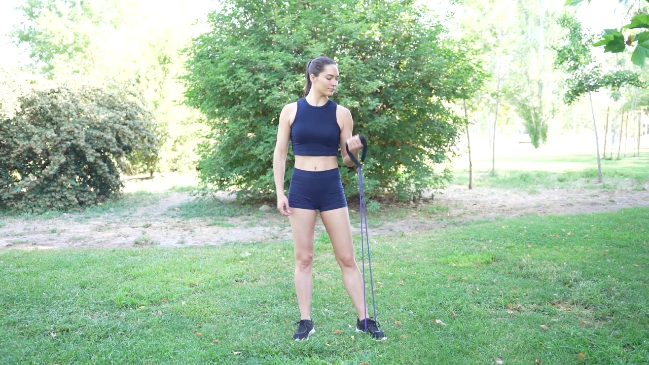 Una donna in abbigliamento sportivo sta facendo esercizi con la fascia di resistenza all'aperto, circondata dal verde.