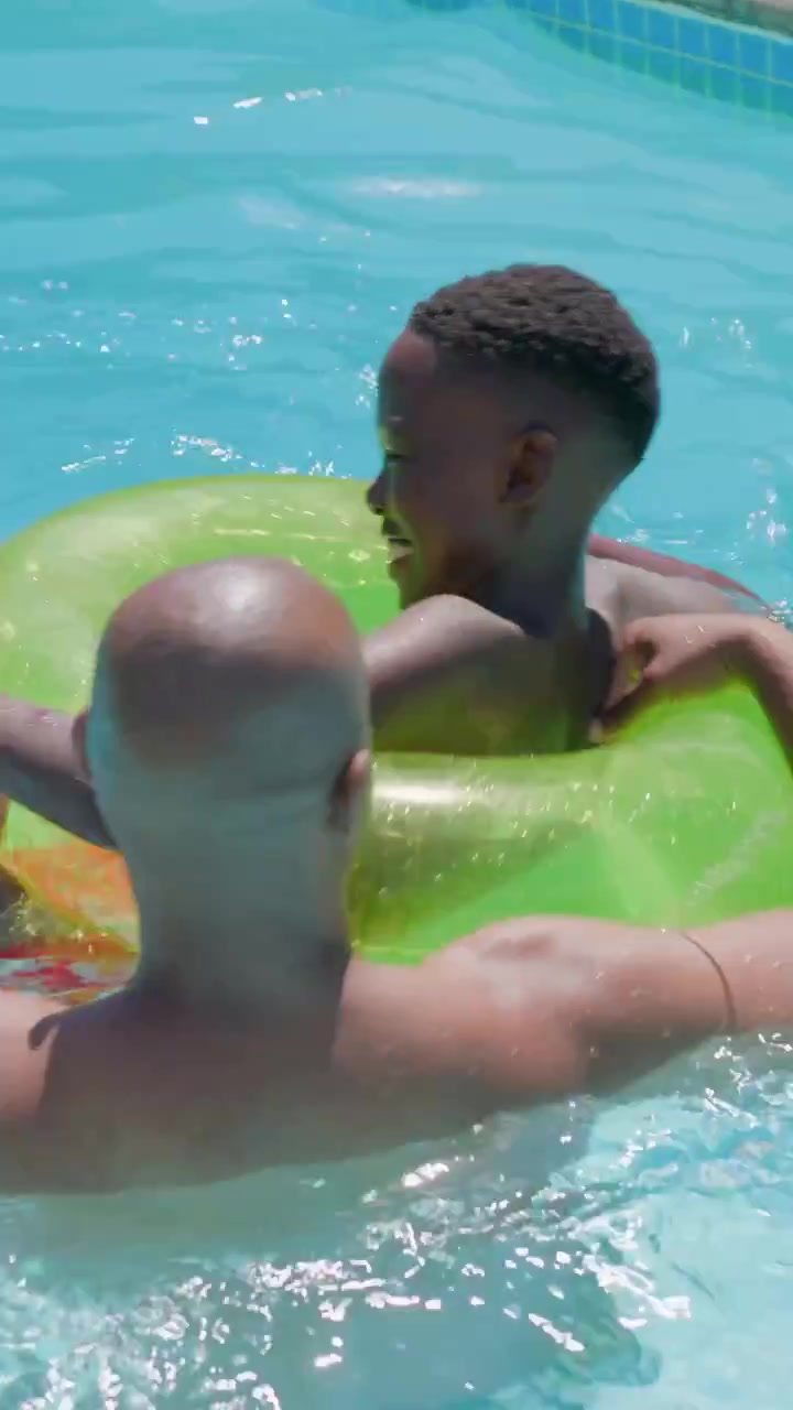 Reaching adult steadying green ring in home pool, youth in red trunks kicking and learning float