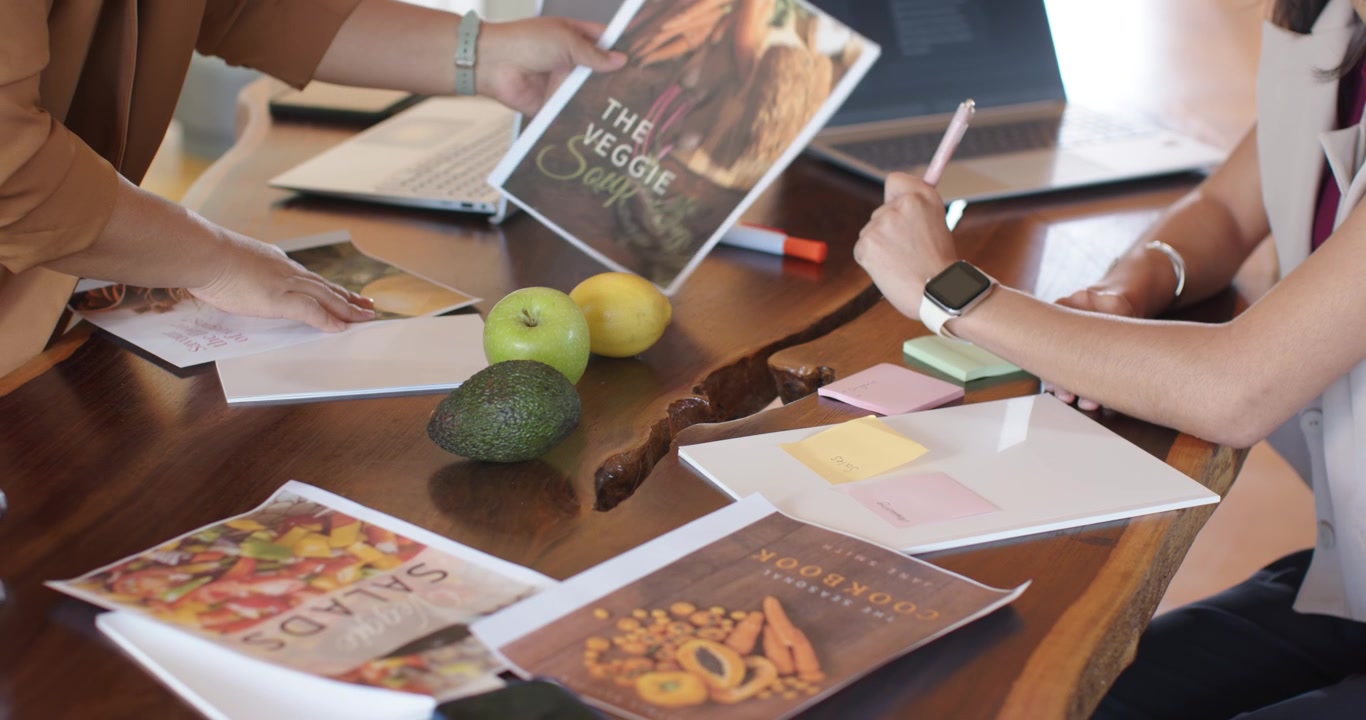 Forskellige kvindelige kolleger styler avocado og æbler, den venstre placerer avocado, den højre skriver layoutnoter.