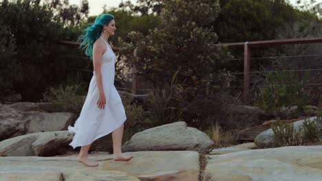 tranquilidad - mujer con vestido blanco y cabello azul camina descalzo sobre rocas en el crepúsculo, slomo