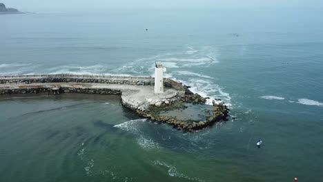 Drone-orbits-around-a-white-small-lighthouse-in-the-edge-of-a-rocky-pier