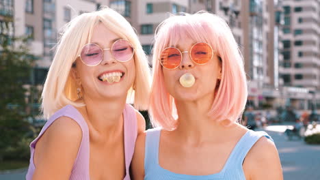 two women with pink and blonde hair, smiling and chewing bubble gum in city