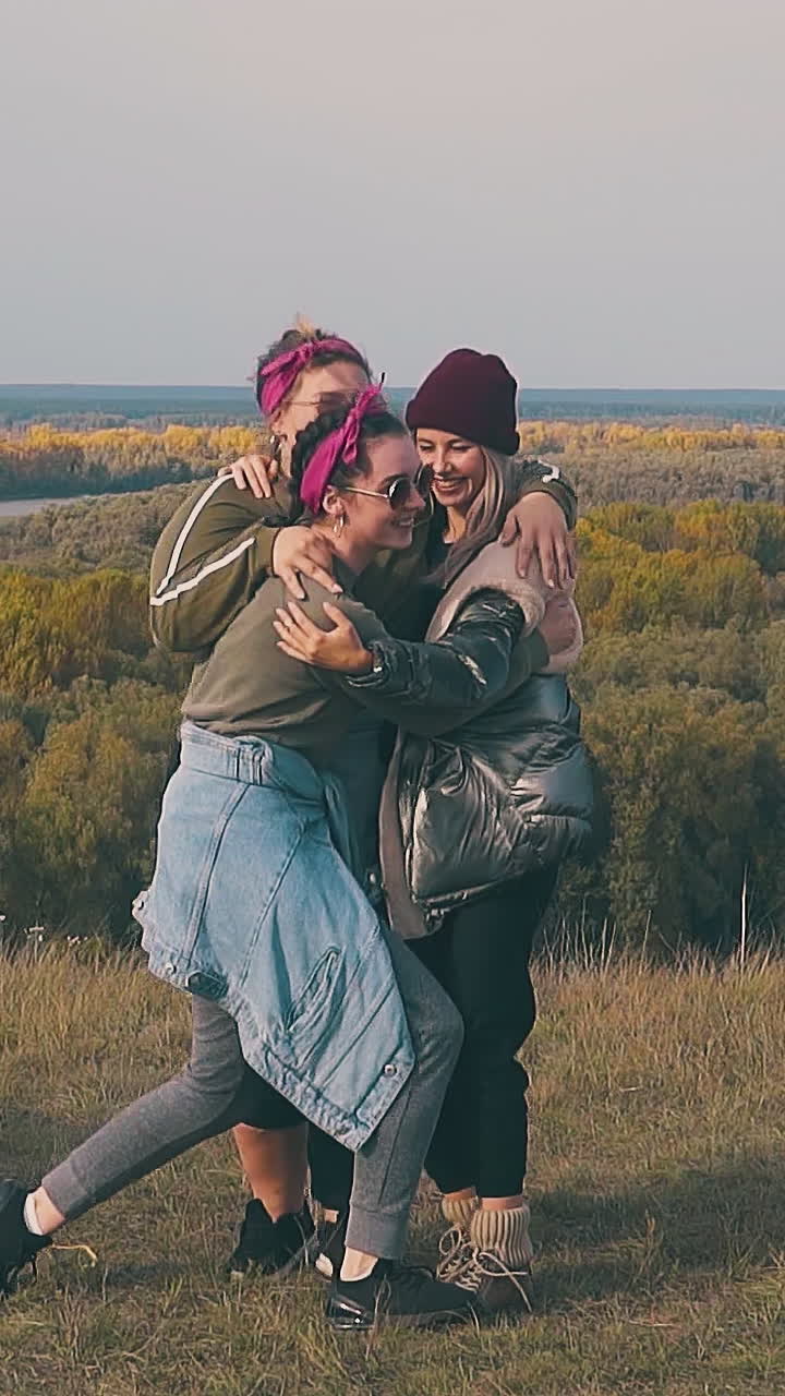 plump young woman hugs girl friends with laughter in campsite against calm river against dense autumn forests slow motion