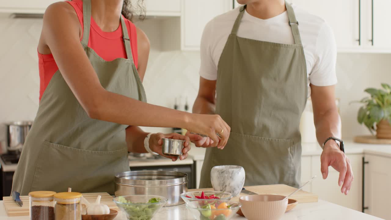 Coppia in grembiule: l'uomo passa il macinino, la donna lo prende e lo sparge nel mortaio in cucina mentre prepara il pasto