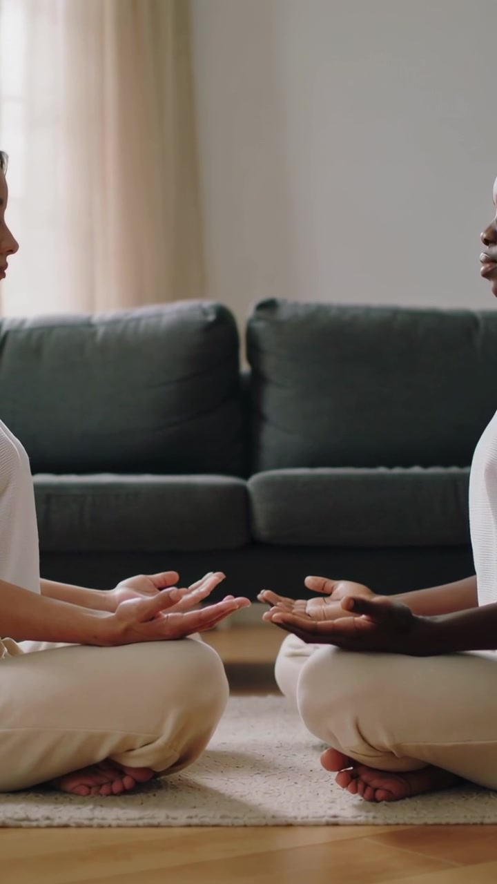 Video verticale: Due donne che sistemano le mani e meditano su un tappeto a casa con un divano verde, indossando pantaloni beige.