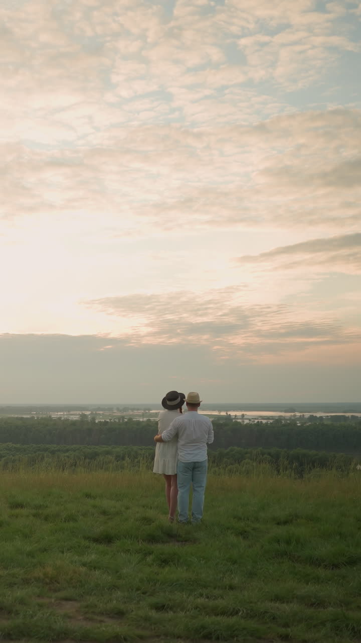 한 부부는 해가 지는 동안 호수를 내려다보는 잔디 언덕 위에 서 있습니다. 남자는 색 셔츠, 모자, 청바지를 입고 여성의 허리를 잡고 있습니다. 그녀는 검은 모자와 색 드레스를 입고 있습니다.