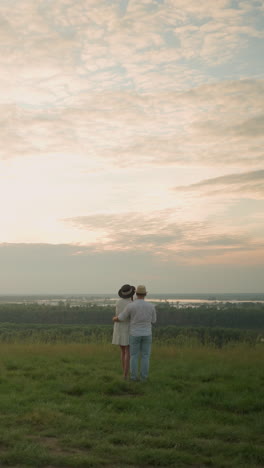 a couple stands on a grassy hill overlooking lake during sunset, shot from behind. the man, in a white shirt, hat, and jeans, holds the woman by the waist. she wears a black hat and a white dress