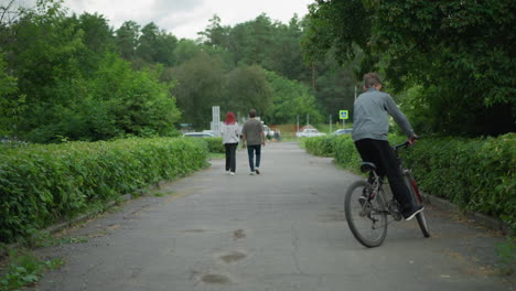 young boy in grey top rides his bicycle in circles on a paved path, two people walk ahead in the distance, with a blurred view of cars in the background, surrounded by lush greenery