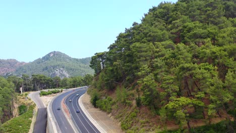 Drone-Aéreo-Volando-Sobre-Una-Colina-De-Montaña-Llena-De-árboles-Al-Lado-De-Una-Carretera-Mientras-Los-Autos-Pasan-En-Un-Día-Soleado-En-Antalya-Turquía