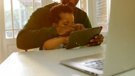 Vista-Frontal-De-Un-Joven-Padre-Negro-Y-Su-Hijo-Usando-Una-Tableta-Digital-En-La-Mesa-Del-Comedor-En-La-Cocina-4k