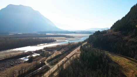 Toma-Aérea-Del-Río-Fraser-Al-Lado-De-La-Autopista-Lougheed-7-En-La-Parte-Baja-Del-Continente-En-Columbia-Británica-En-Canadá,-Escena-Brillante-Con-Montañas-Nubladas-En-Otoño-Con-Autos-Conduciendo-Por-La-Carretera