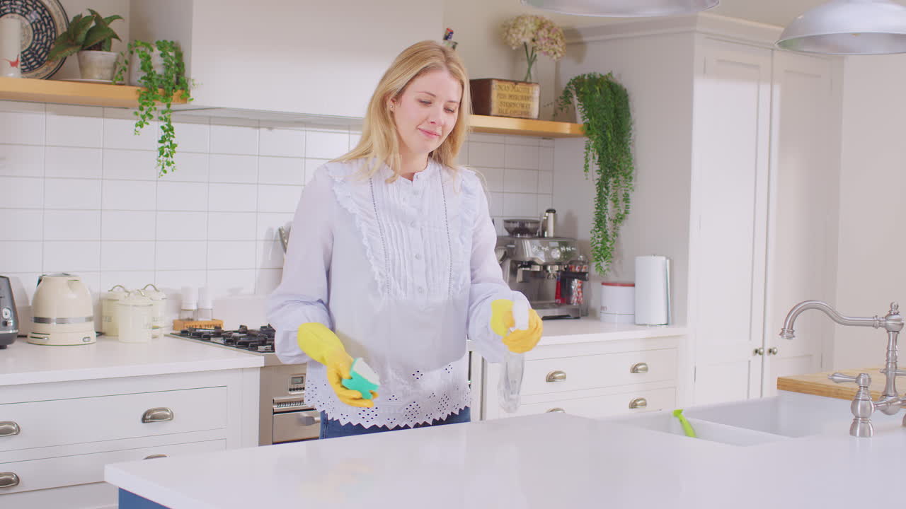 Panning shot of woman at home in kitchen wearing rubber gloves cleaning down work surface using cleaning spray - shot in slow motion