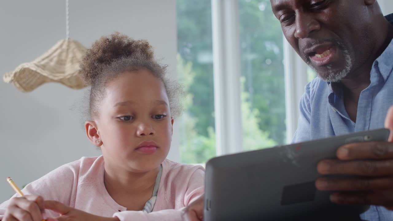 Nonno che aiuta la nipote con la scuola a casa, seduti al tavolo con un tablet.