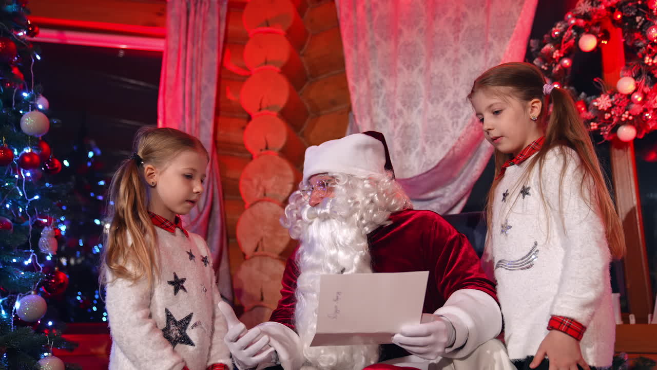 Children standing near Santa and talking to him. Kids sharing pictures and their Christmas dreams with Santa Claus. Santa asking girls about their wishes for holiday.