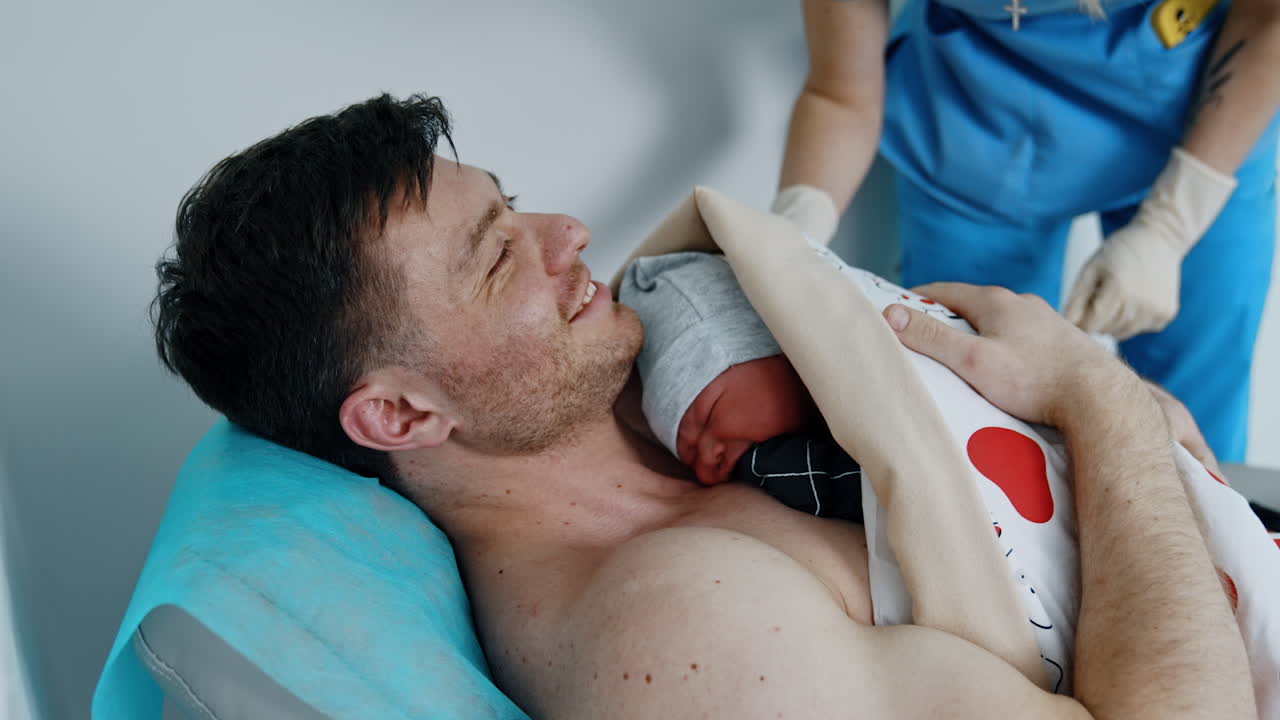 Newborn child is put on the bare chest of his father. Happy dad is smiling. Nurse helping to cover the infant.