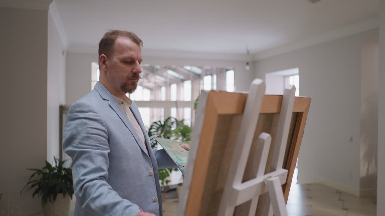 아티스트는 스튜디오에서 스에서 일합니다. 사려 깊은 디자이너는 미술 작업실에서 큰 캔버스에 예술 작품을 그립니다. 재을 입은 수염이있는 남자는 걸작에 색을 적용합니다.