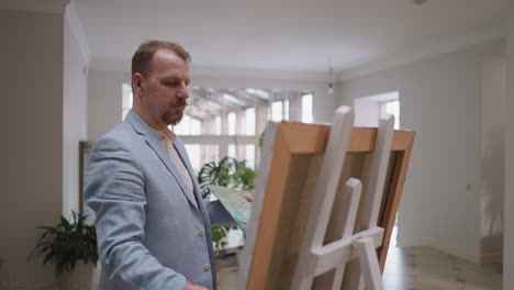 el artista trabaja en el caballete en el estudio. el diseñador pensativo dibuja obras de arte en grandes lienzos en el taller de arte. el hombre barbudo en chaqueta aplica color a la obra maestra