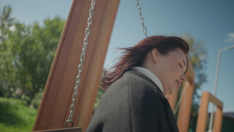 close-up of woman swinging outdoors, her hair gently swaying with breeze, holding chain, set against a sunny park with blurred people and lush greenery