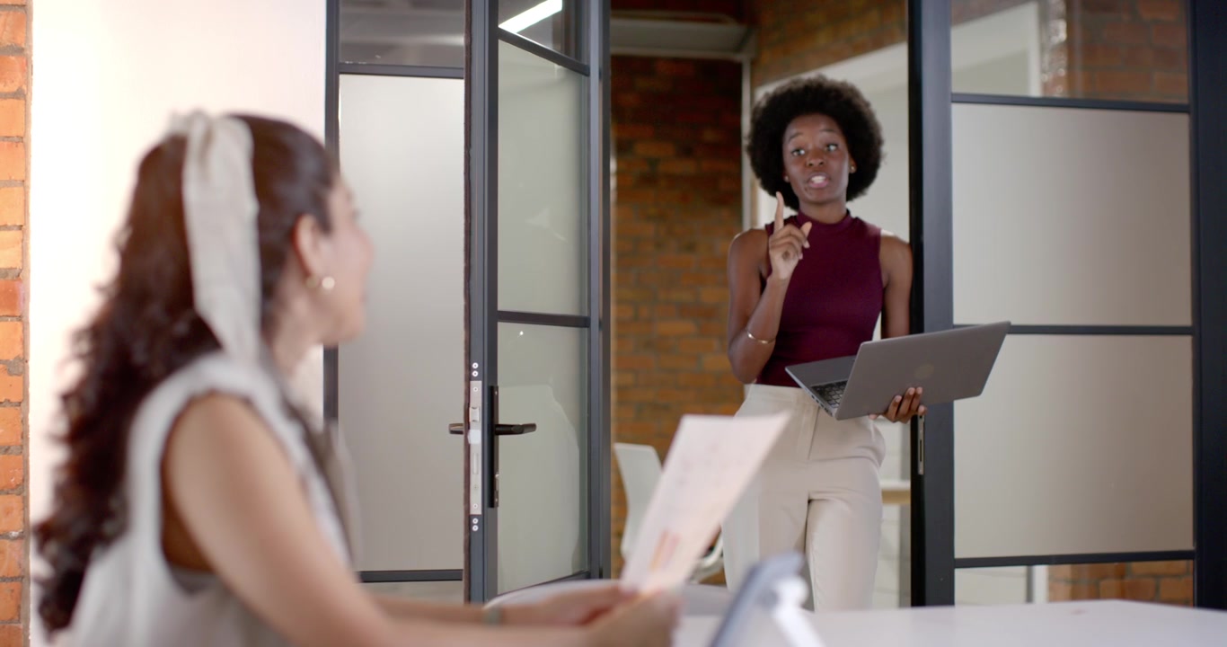 Colegas de trabalho (mulheres) revisando gráficos em uma mesa de reunião, enquanto uma mulher africana em pé fala com um laptop.