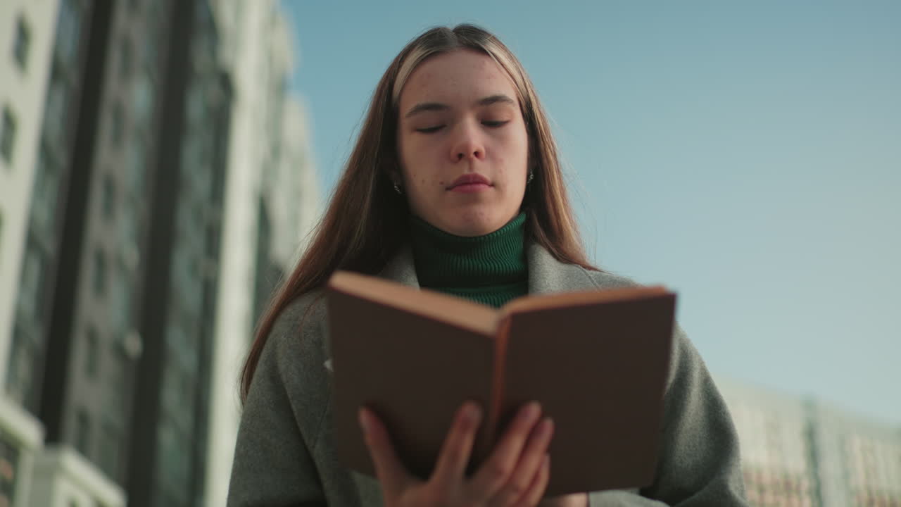 Mulher de casaco cinza caminha ao ar livre lendo um livro com expressão concentrada, edifícios residenciais modernos borrados ao fundo, capturando estilo de vida, aprendizado e atividade reflexiva na cidade.