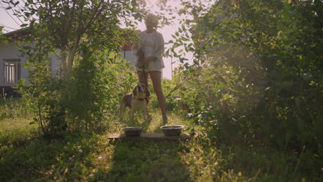 woman standing with her dog between her legs holding leash as dog barks, with two metal bowls placed on plank in front of them, surrounded by lush greenery, while sunlight filters through from behind