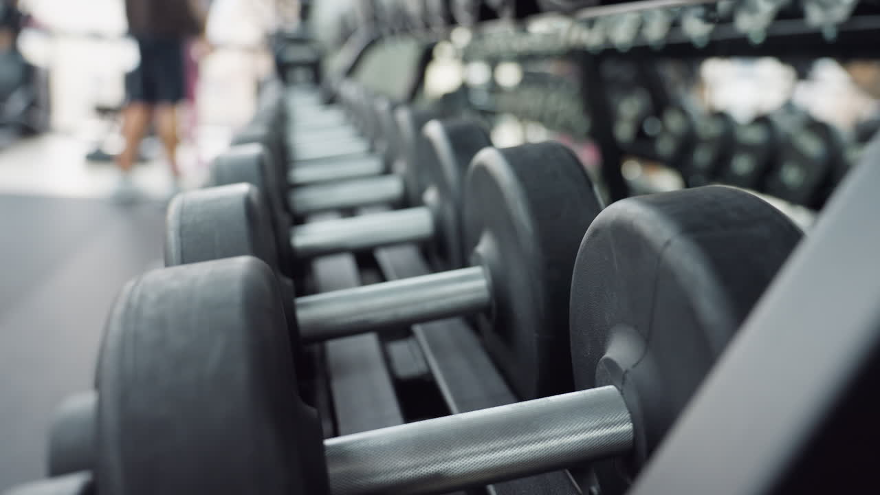 Dumbbells lined up on gym rack with blurred athletes in background, highlighting sleek weight design, textured handles, modern fitness environment, focus on equipment readiness for strength