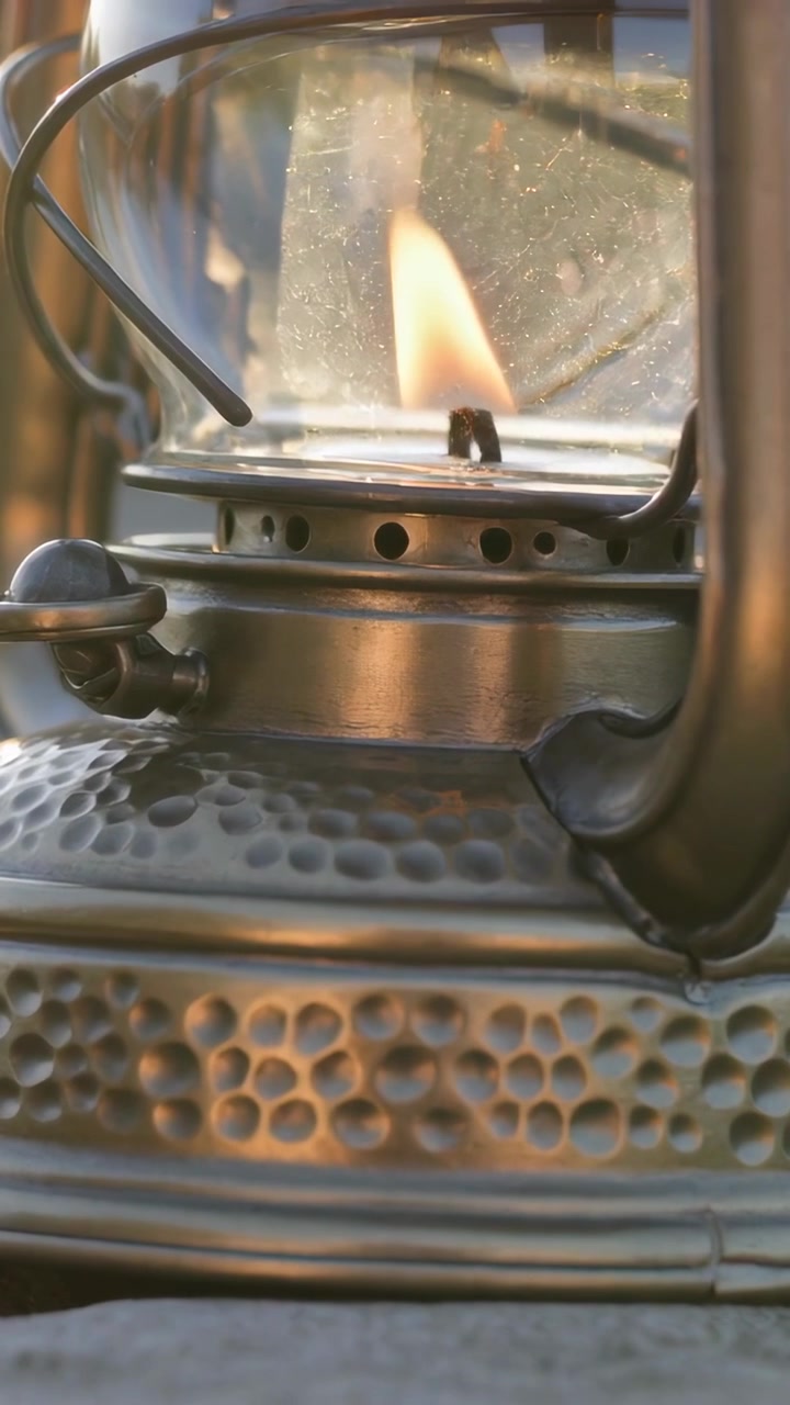 Video vertical: Linterna encendida mostrando la llama de la mecha inclinada por la brisa en una mesa exterior, con inclinación del encuadre.