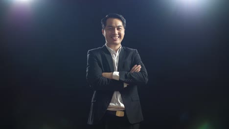 smiling asian speaker man in business suit crossing his arms while standing in the black screen studio