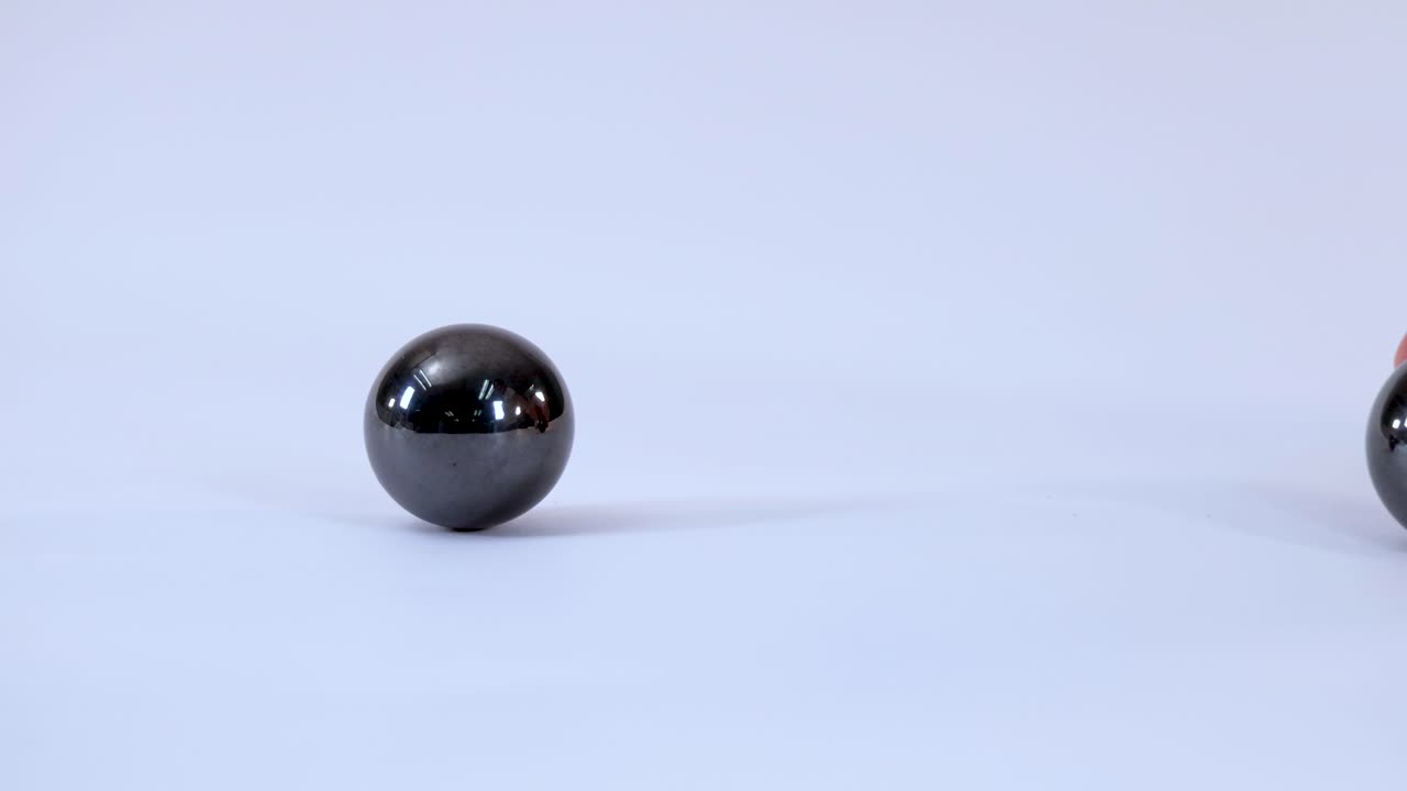 A hand moves a shiny magnetic ball toward another, demonstrating attraction on white background