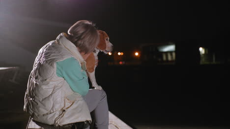 instructor holds her dog firmly on lap during calm night outdoors under gentle spotlight, both sharing warm, secure connection filled with love, and peaceful companionship on empty wooden benches