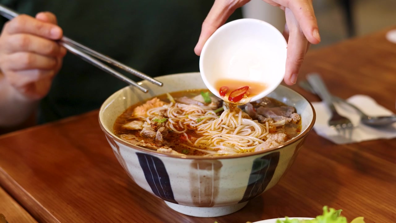 Primo piano di mani che versano salsa di pesce in una ciotola di ceramica di noodles vietnamiti tradizionali
