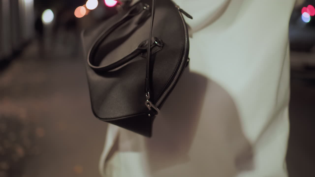Close-up back view of stylish lady carrying elegant black designer handbag, white coat flowing as she strolls under city lights at night with soft bokeh background