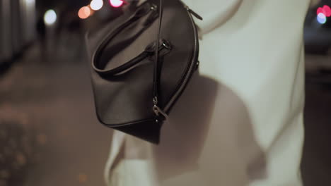 close-up back view of stylish lady carrying elegant black designer handbag, white coat flowing as she strolls under city lights at night with soft bokeh background