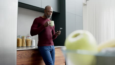 Happy-mature-biracial-man-drinking-coffee-and-using-smartphone-in-kitchen,-slow-motion