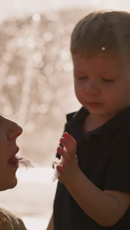cute little boy feeds mother with spun sugar against fountain at sunset closeup slow motion. child treats woman with dessert. happy motherhood