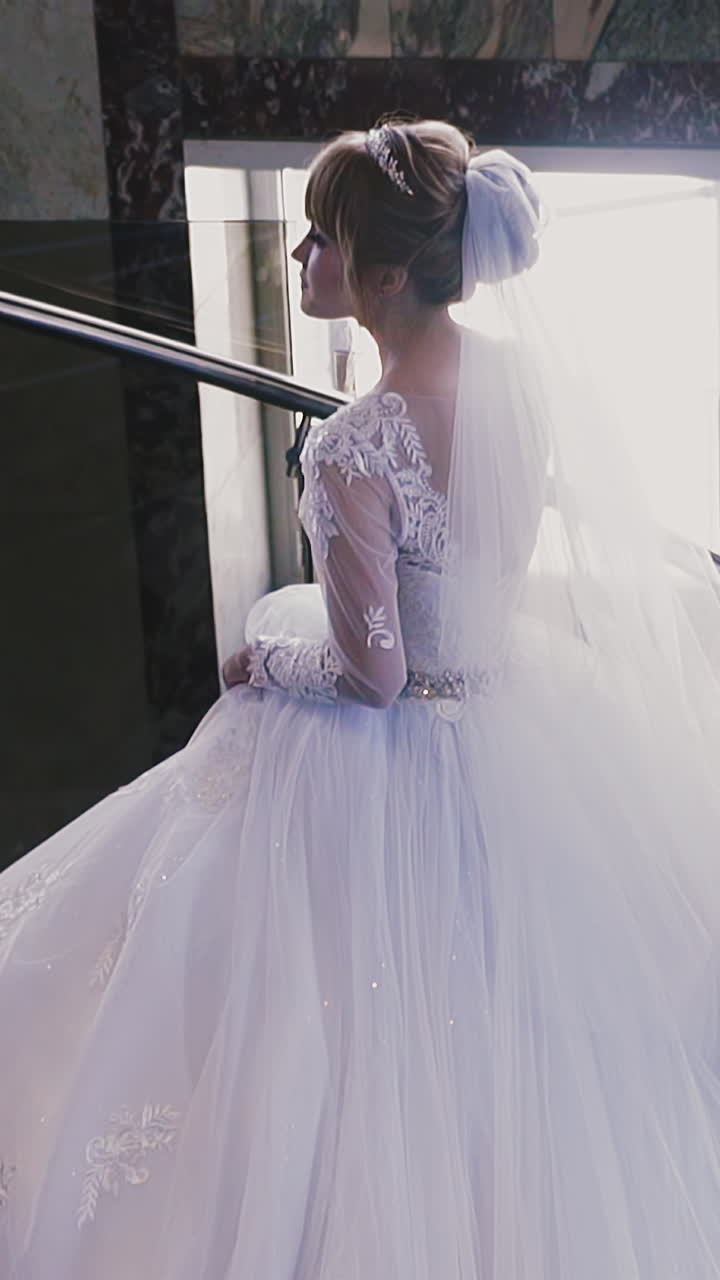 Slow motion: elegant young bride with white veil and gorgeous lacy wedding dress goes up stairs leaning on metal railing in spacious hall closeup
