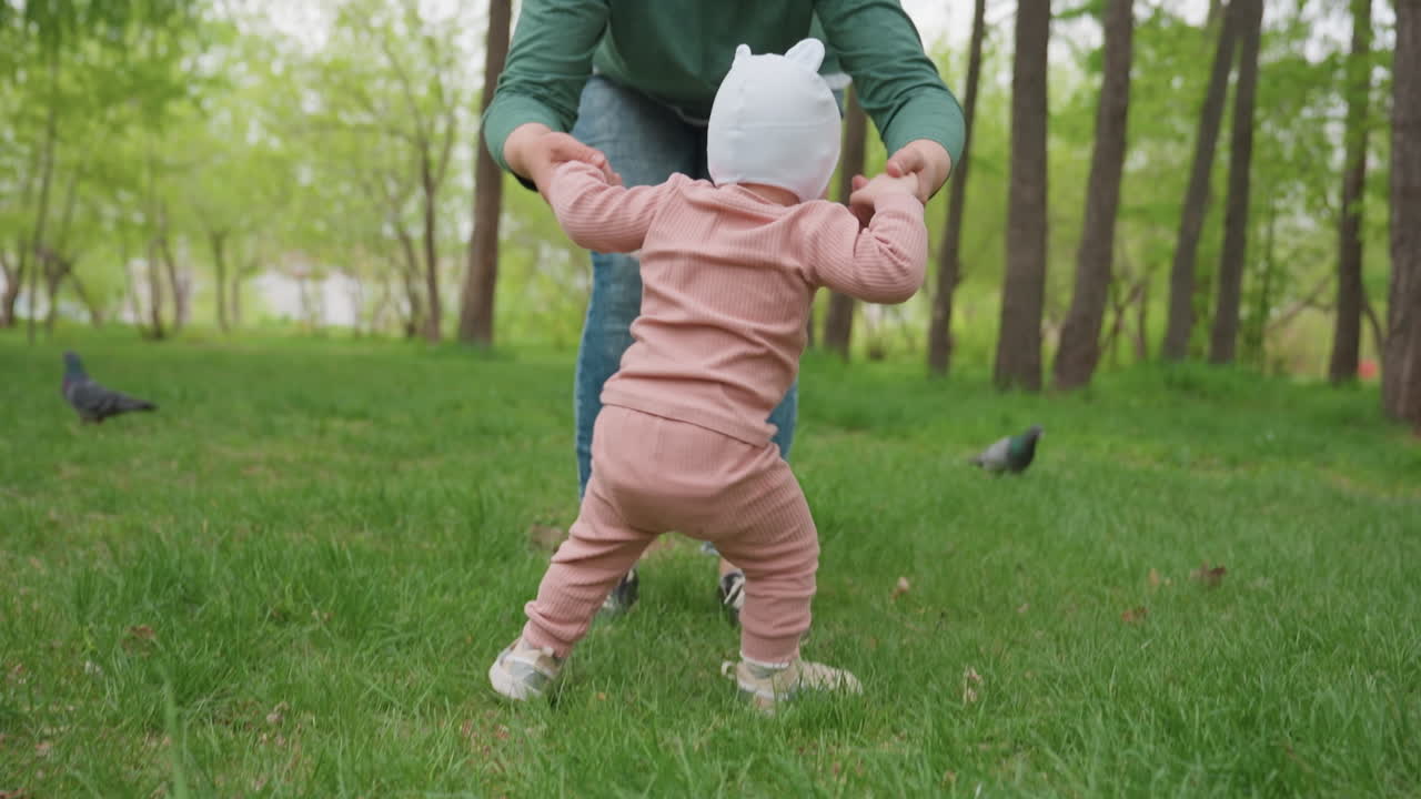 Baby Learns To Walk Outdoors, Young Child Begins To Walk With Gentle Encouragement Outdoors In Nature, Little Baby Takes Initial Steps On Soft Grass As Caring Adult Guides And Encourages