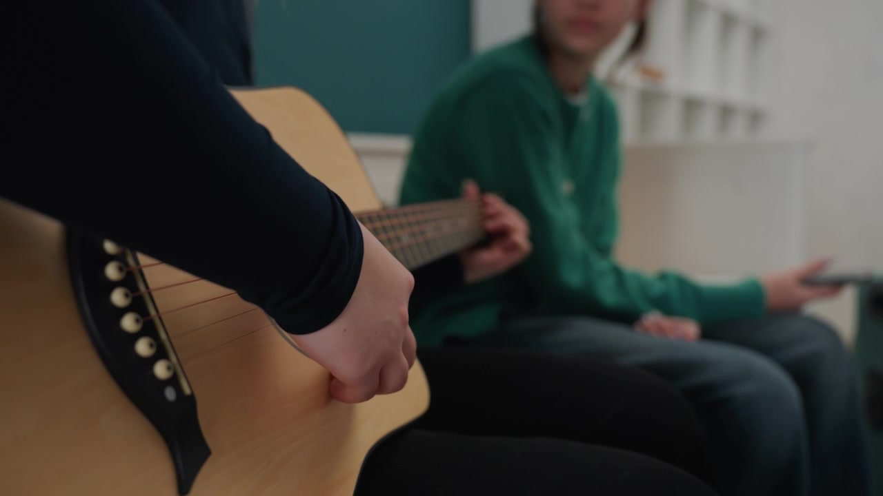 Dos personas en el sofá tocando la guitarra en una sesión de improvisación en apartamento, uno guiando al otro, ambiente de lección informal, detalle de suéter verde, primeros planos de la mano rasgueando cerca de la boca de la guitarra, atmósfera relajada, en movimiento.
