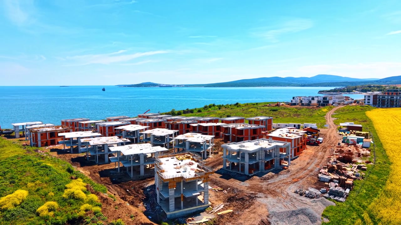 Coastal residential development project under construction near blue sea water. Aerial view of concrete cottage frameworks on a green hill overlooking the ocean.