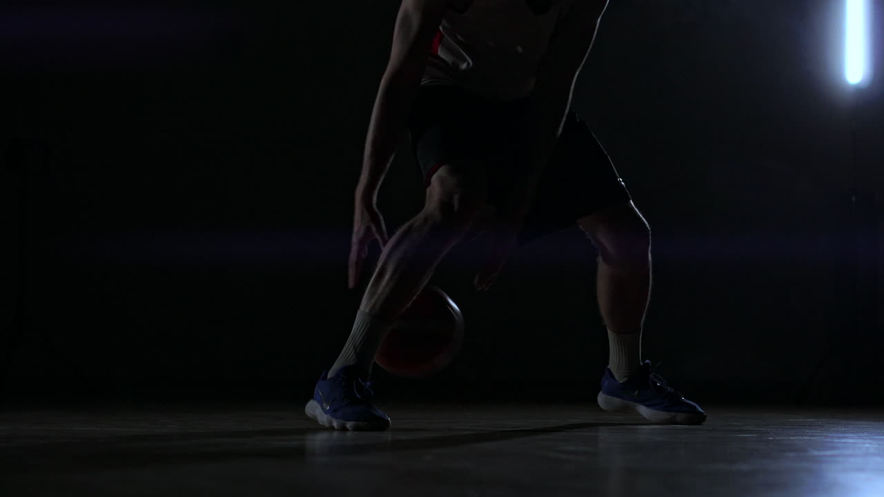 A man with a basketball on a dark basketball court against the backdrop of a basketball ring in the smoke shows dribbling skills illuminated by three lanterns in backlight