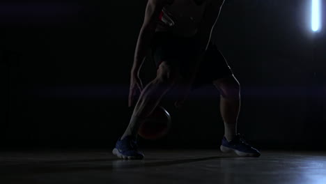 A-man-with-a-basketball-on-a-dark-basketball-court-against-the-backdrop-of-a-basketball-ring-in-the-smoke-shows-dribbling-skills-illuminated-by-three-lanterns-in-backlight