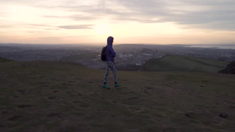 circling parallax shot of girl walking on top of the arthurs seat mountain on the hiking trail in evening, dusk, blue hour with cityscape of edinburgh in the background