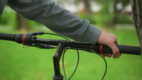 vista parcial de una bicicleta estacionada con la mano en ropa gris sosteniendo y ajustando el manillar, comprobando la funcionalidad de los frenos, el fondo está borroso, mostrando un exuberante parque verde