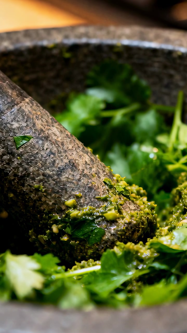 Vertical video: Pressing stone mortar, pestle crushing green herbs in kitchen, releasing oils