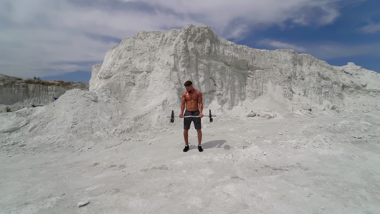 Bodybuilder is doing his workout in amazing white mountains. Muscular man only in shorts training with heavy barbell outdoors on the natural mountain background.