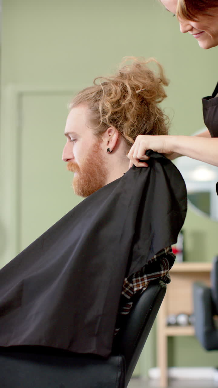 Vertical video of caucasian female hairdresser putting cape on client at hair salon, in slow motion