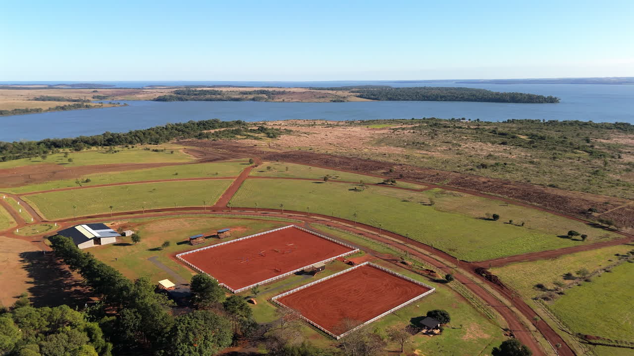 Drone panning shot shows equestrian field with training arenas, buildings, and open farmland near a wide river, dry terrain and green patches under blue sky, real time, drone pan motion