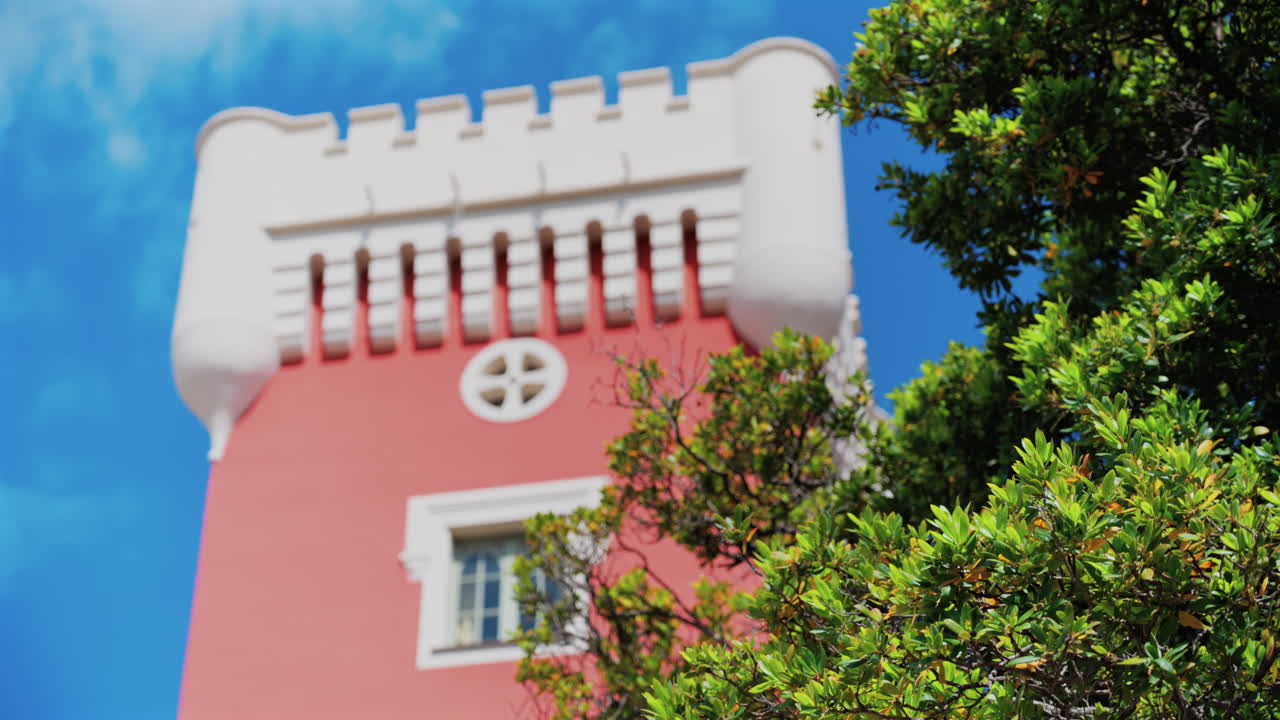 Green tree moving in the wind near the red tower of the Cremat Castle Winery over blue sky