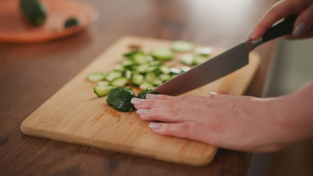 Hand with polished nails resting on cutting board while slicing fresh cucumber into pieces, showing detailed view of knife action and fresh vegetables prepared on kitchen counter